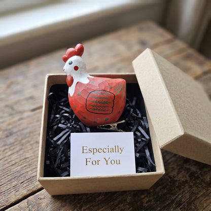 A hand-painted wooden rooster ornament in red, white, and black resting in an open kraft gift box filled with black shredded paper, featuring a small card reading "Especially For You" placed in the foreground.