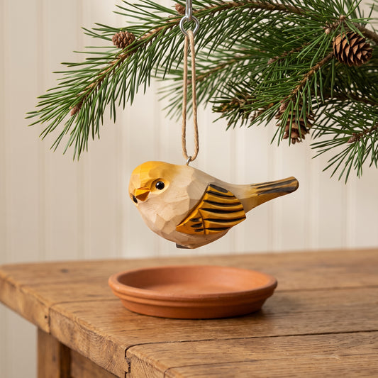 A carved wooden bird ornament with yellow, beige, and black details hangs from a pine branch by a twine loop, positioned above a terracotta saucer on a rustic wooden table.