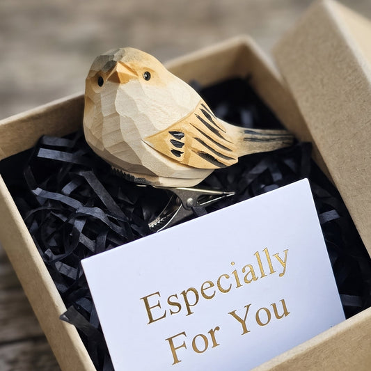 A hand-carved wooden chubby bird clip-on ornament resting inside an open kraft paper gift box filled with black shredded paper, featuring an orange-toned head, a cream throat, a beige body with black wing accents, and a white card reading "Especially For You" placed in the foreground on a rustic wooden surface.