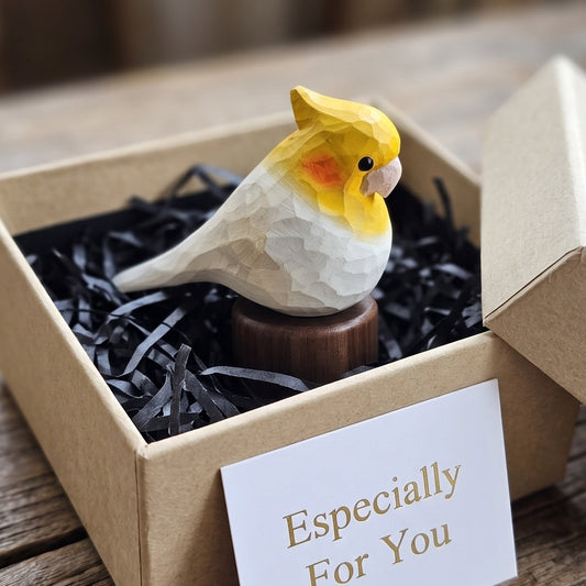A small carved Cockatiel figurine in yellow, white, and orange sits in a brown gift box filled with black shredded paper, with a card reading 'Especially For You' on a rustic wooden table.