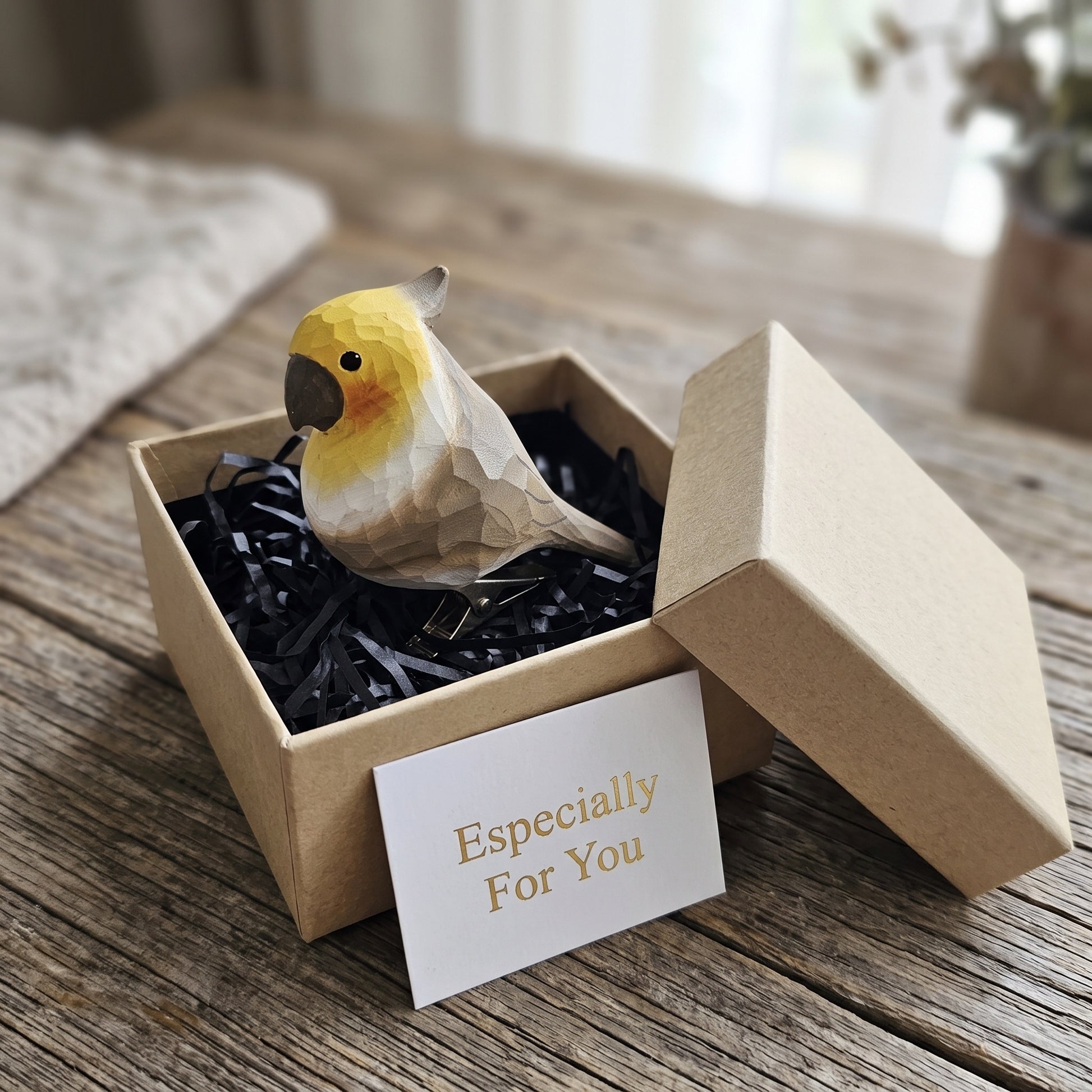 Hand-carved gray cockatiel wooden ornament sitting in an open kraft gift box with black crinkle paper and an "Especially For You" card.
