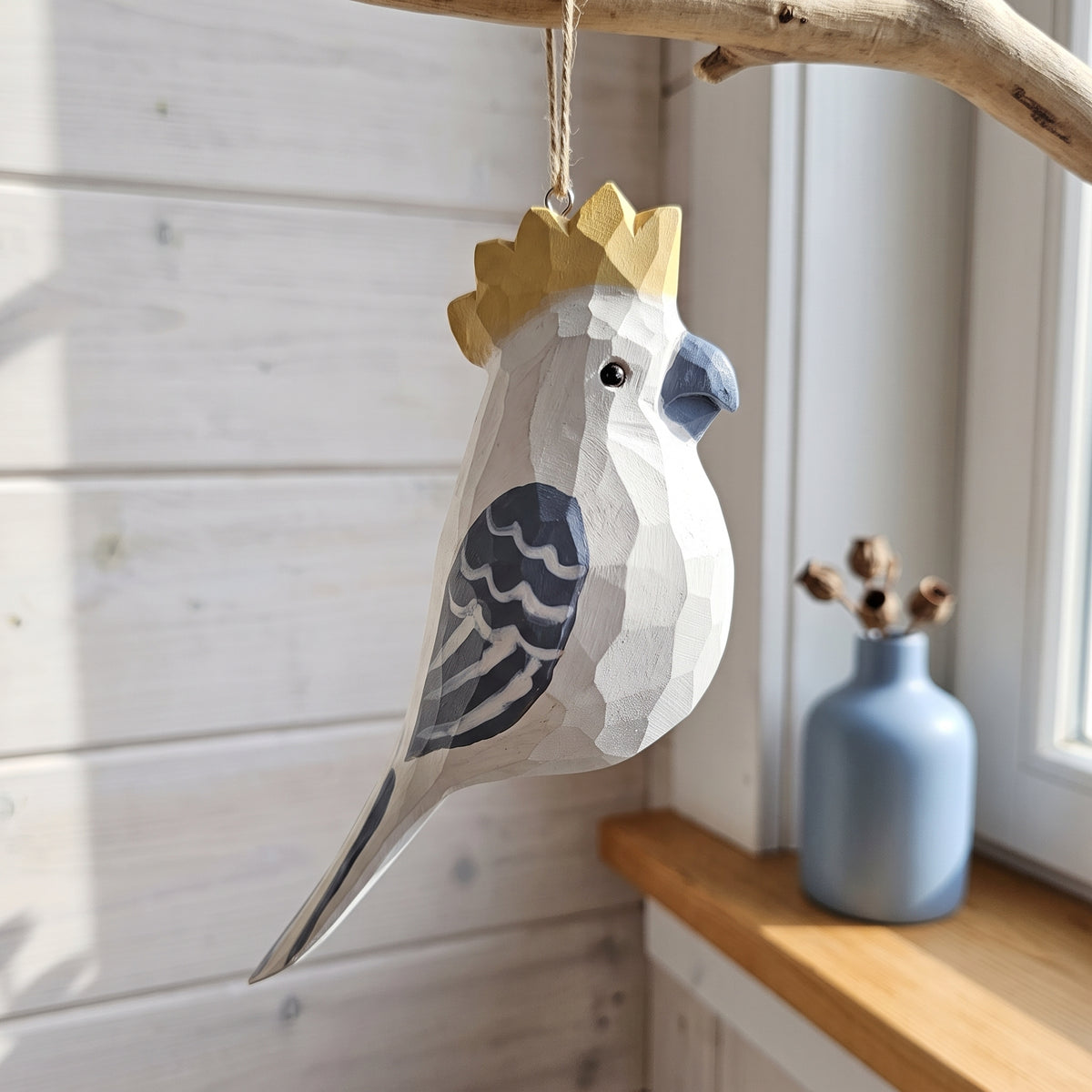 A geometric, faceted wooden cockatoo hanging ornament with a gold crown, hanging from a rustic branch in a sunlit room next to a blue vase.