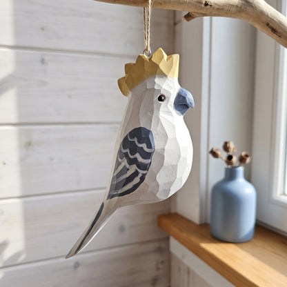 A geometric, faceted wooden cockatoo hanging ornament with a gold crown, hanging from a rustic branch in a sunlit room next to a blue vase.