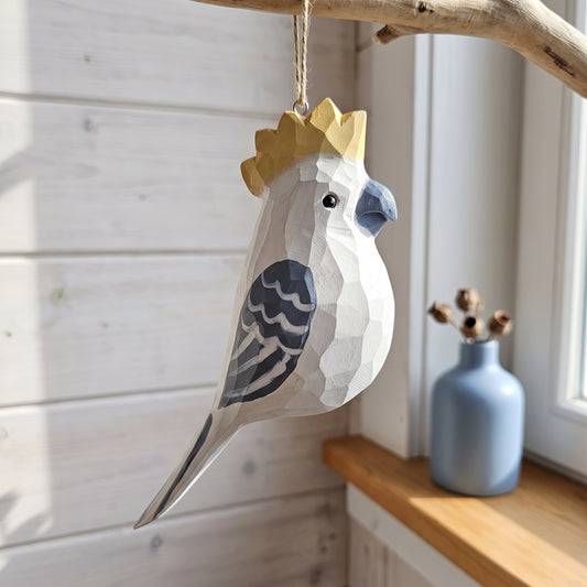 A geometric, faceted wooden cockatoo hanging ornament with a gold crown, hanging from a rustic branch in a sunlit room next to a blue vase.
