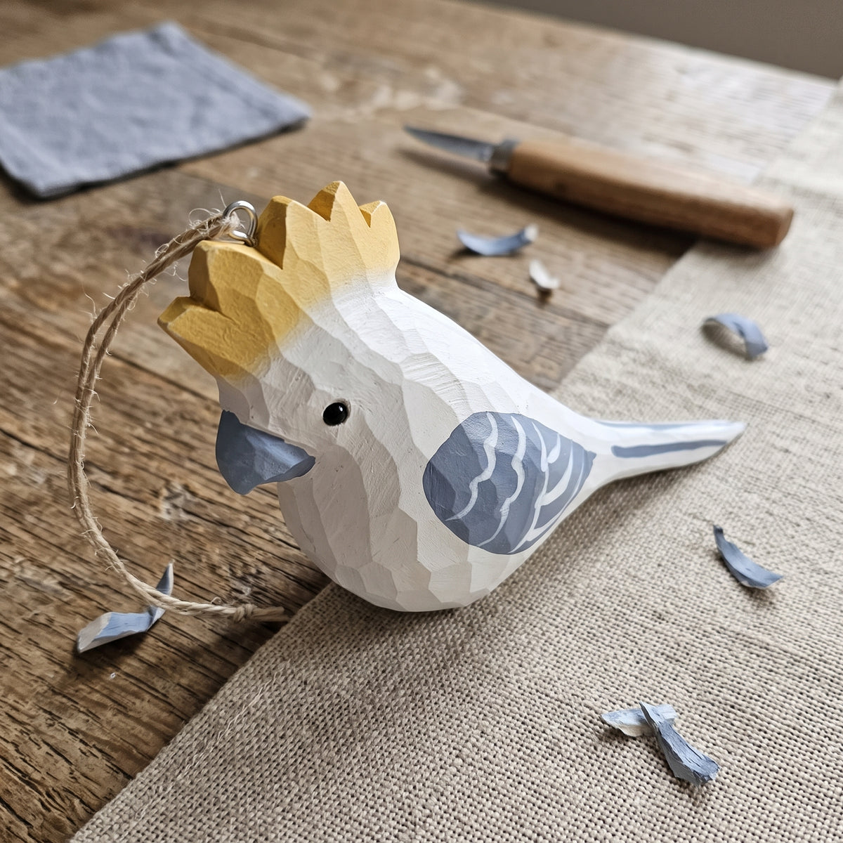 A handcrafted wooden Cockatoo ornament with a yellow and white gradient crest, blue beak, and blue patterned wing, resting on a burlap-covered wooden table with carving tools and wood shavings nearby.
