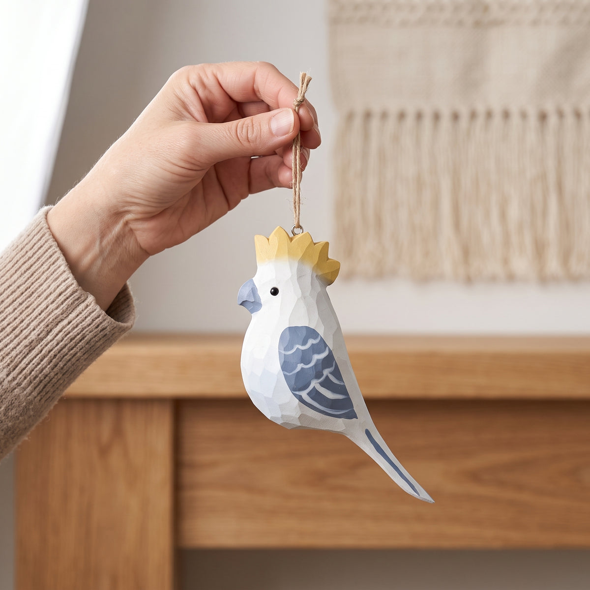 A hand holding a stylized, low-poly paper cockatoo ornament with a yellow crown, blue wing, and blue beak, set against a warm, softly blurred home background.