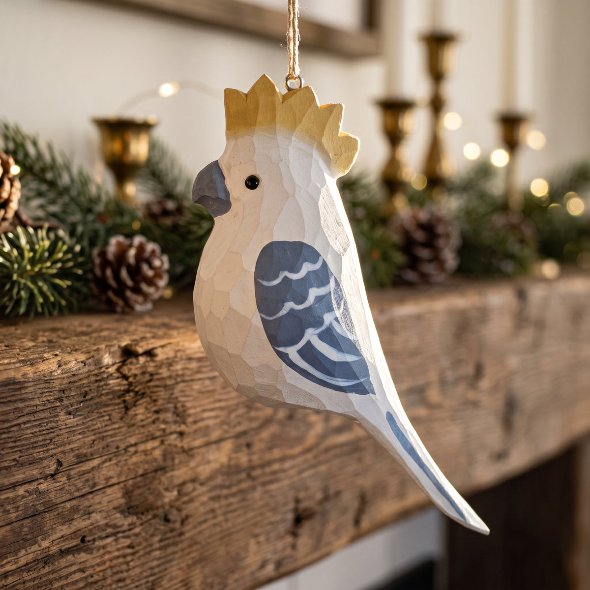 A geometric, faceted wooden cockatoo bird ornament hanging from a string in a cozy holiday setting with a rustic wooden mantel, pinecones, and blurred candles in the background.