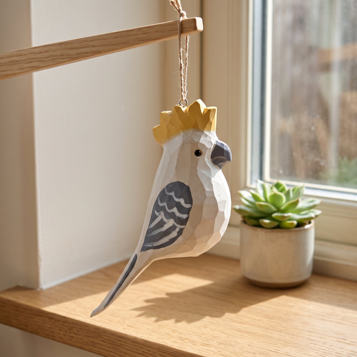 A low-poly wooden Cockatoo ornament with a gold crown hangs from a wooden rod in a sunlit room next to a small potted succulent.