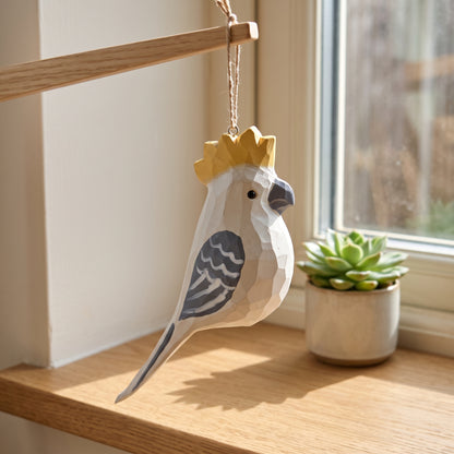 A low-poly wooden Cockatoo ornament with a gold crown hangs from a wooden rod in a sunlit room next to a small potted succulent.