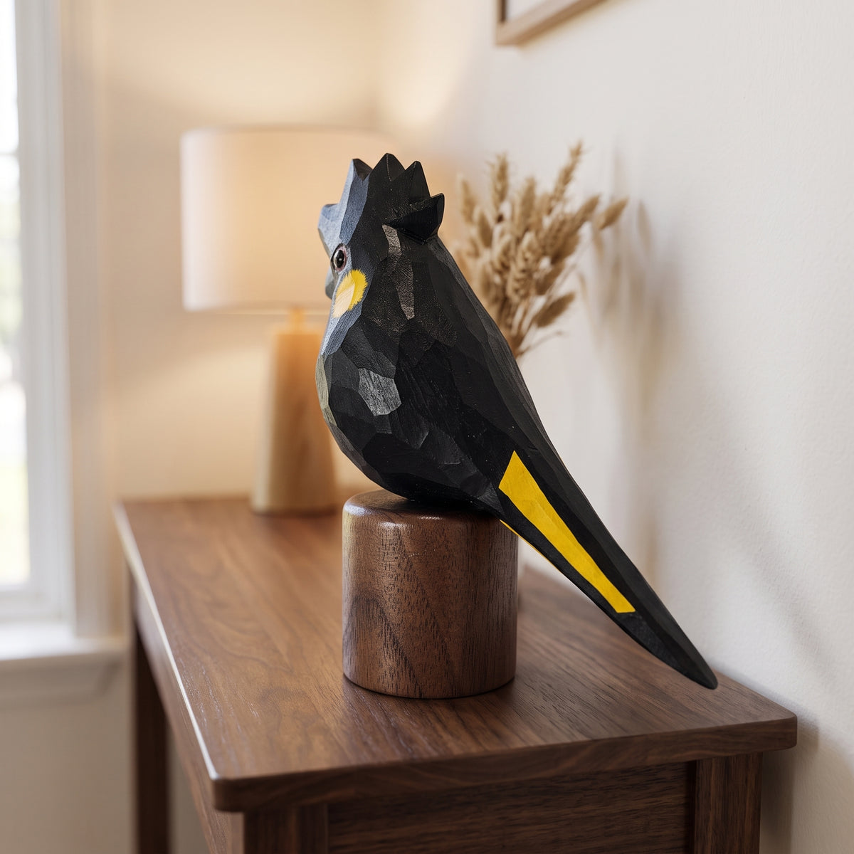Wooden Yellow-tailed Black Cockatoo statuette on round walnut base placed on a tabletop next to a lamp