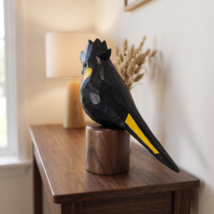 Wooden Yellow-tailed Black Cockatoo statuette on round walnut base placed on a tabletop next to a lamp