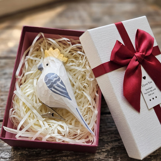 A white Cockatoo bird-shaped hair clip with blue-gray wings and a small yellow crown, displayed inside an open burgundy gift box with shredded paper next to a white gift box with a red ribbon and bow.