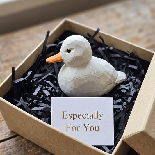 A white geometric Coleduck figurine with an orange beak resting in black shredded paper inside a kraft gift box, with a card reading 'Especially For You' placed in front.