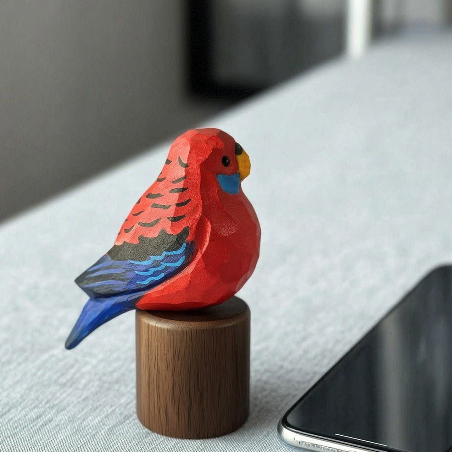 A small carved wooden Crimson rosella figurine in bright red, blue, and black sits on a cylindrical wooden base next to a smartphone on a light gray fabric-covered surface.
