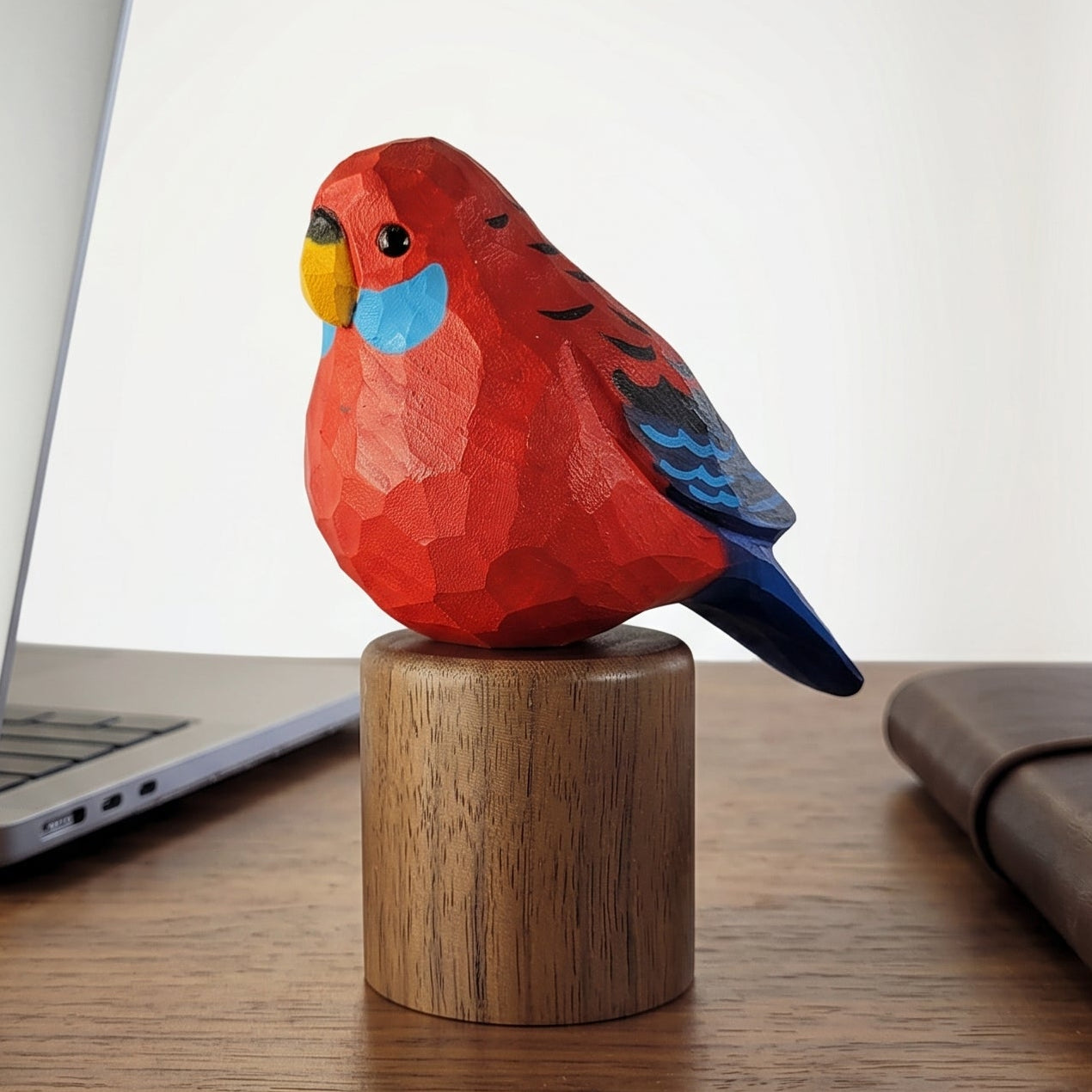 A small hand-painted wooden Crimson rosella figurine in red, blue, and yellow sits on a cylindrical wooden base on a wooden desk, with a laptop, notebook, and window in the background.