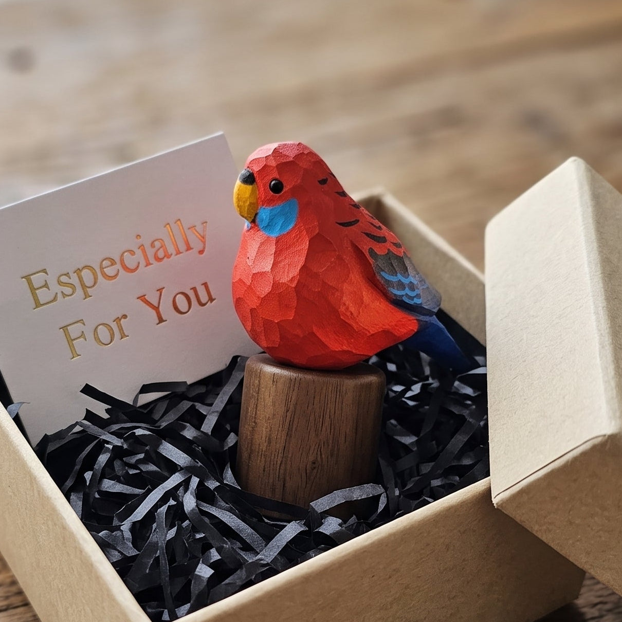 A small Crimson rosella figurine with blue and yellow accents sits on a wooden stand inside a kraft gift box with black shredded paper and a card that reads 'Especially For You,' placed on a wooden table.