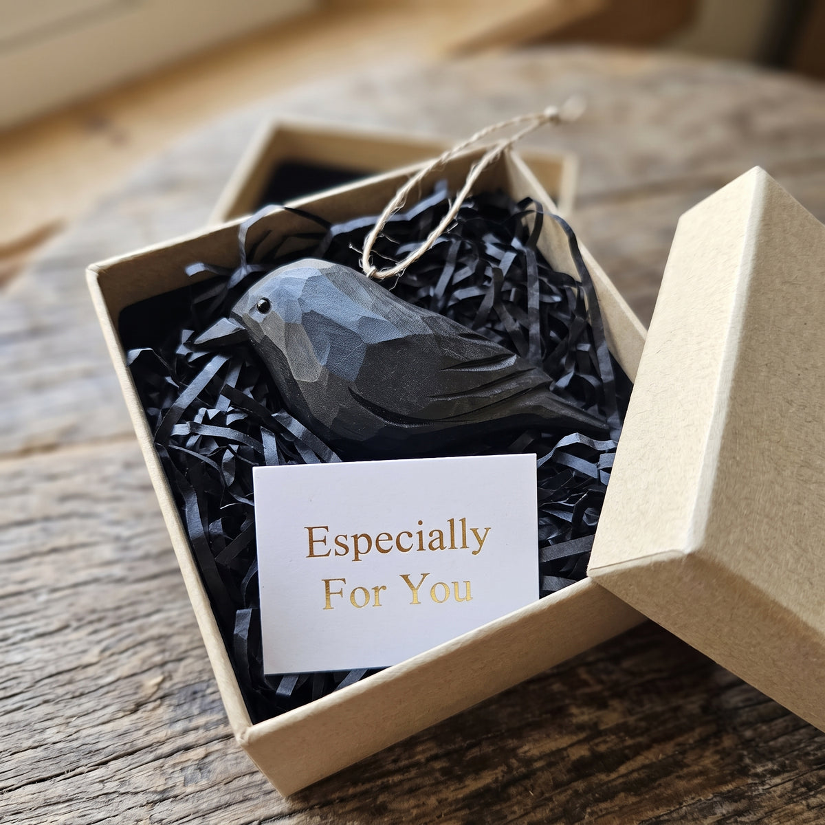 A faceted Crow ornament nestled in black shredded paper inside a kraft gift box, with a small card reading 'Especially For You' and the box lid leaning beside it on a rustic wooden surface.