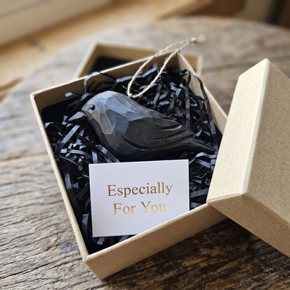 A faceted Crow ornament nestled in black shredded paper inside a kraft gift box, with a small card reading 'Especially For You' and the box lid leaning beside it on a rustic wooden surface.