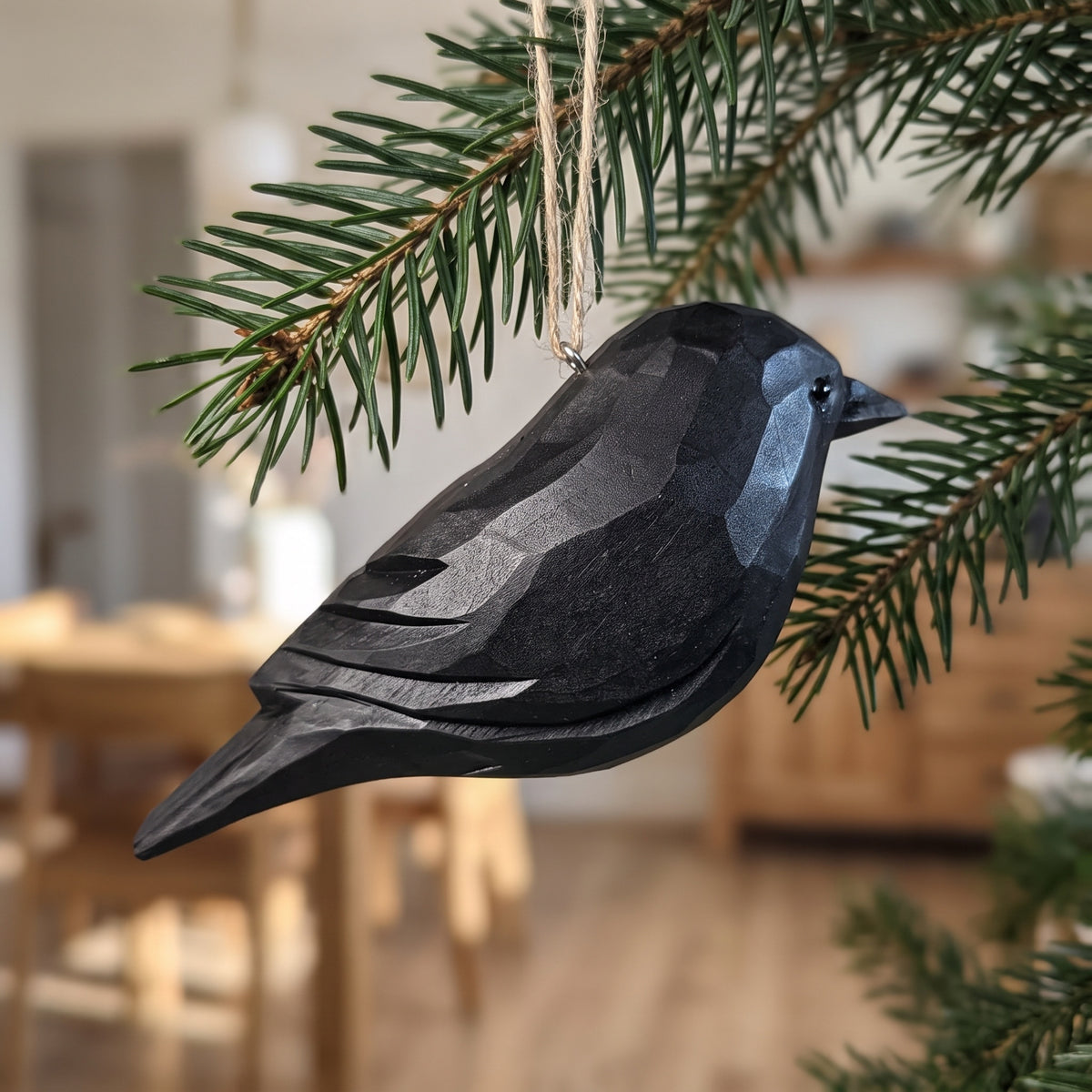 A dark gray wooden Crow Christmas ornament with a faceted geometric design, hanging from a natural jute string on a pine tree branch in a softly blurred indoor setting.