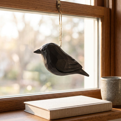 A dark gray geometric Crow ornament hanging from a window by a string, with a book and ceramic mug on the windowsill in a softly lit room.