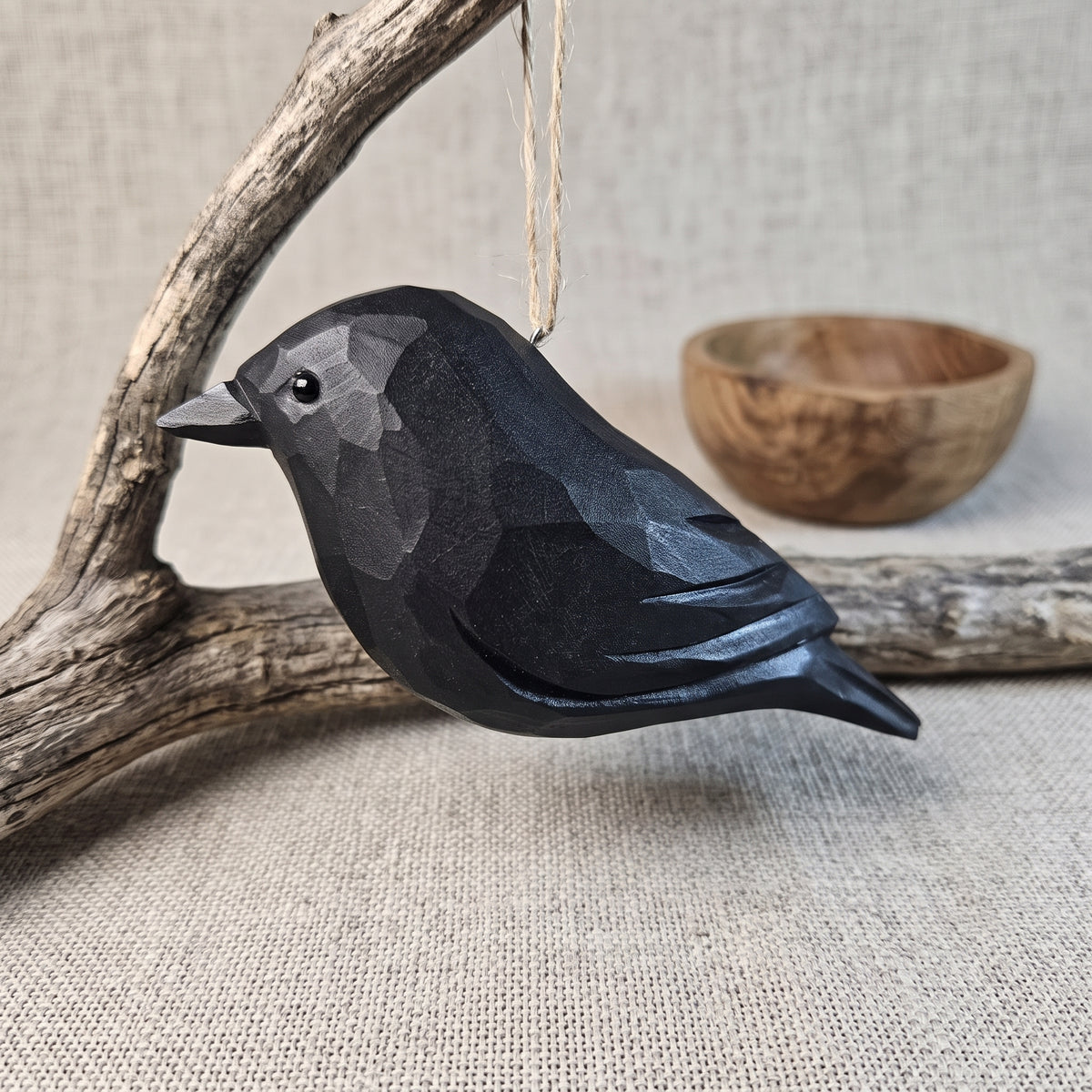 A black faceted Crow ornament hanging from a string, posed on a piece of driftwood with a small wooden bowl in the background on a linen surface.