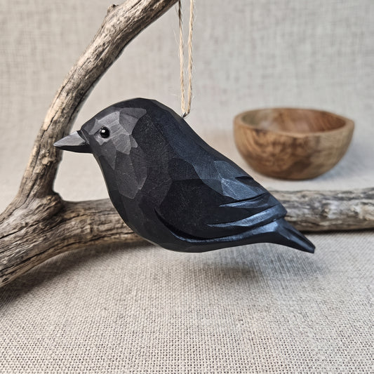 A black faceted Crow ornament hanging from a string, posed on a piece of driftwood with a small wooden bowl in the background on a linen surface.