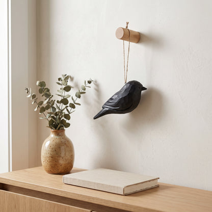 A black Crow wall hanging suspended from a small wooden rod on a cream wall, styled above a light wood console table with a vase of eucalyptus and a book.