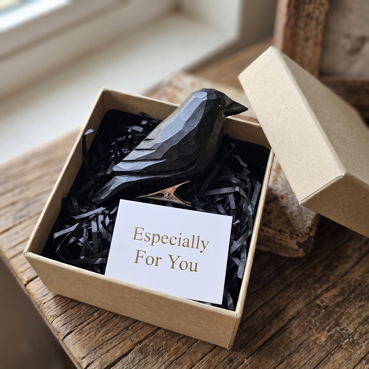 A black Crow-shaped hair claw clip nestled in black shredded paper inside a kraft gift box, with a white card reading 'Especially For You' and an open kraft box beside it on a rustic wooden surface.