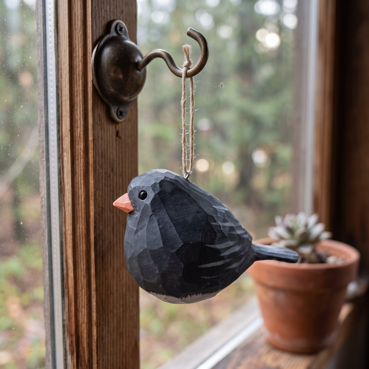 A faceted dark gray Dark-eyed Junco ornament with a small orange beak hangs from a metal hook mounted on a wooden window frame, with a blurred outdoor scene and a potted succulent in the background.
