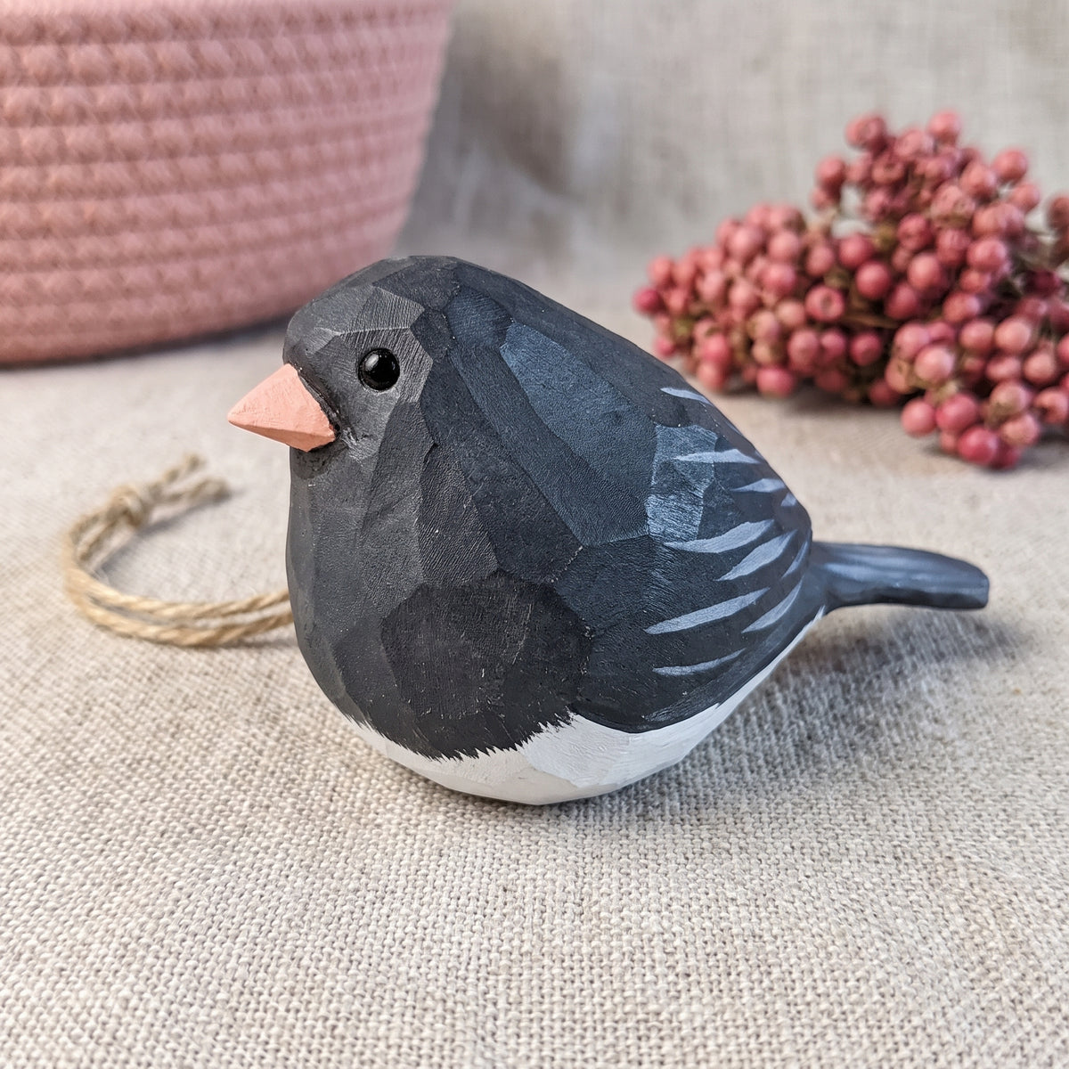 A small Dark-eyed Junco-shaped decorative ornament with a faceted geometric design, painted in charcoal gray, white, and a peach beak, resting on a beige fabric surface with a pink woven basket and pink berries in the background.