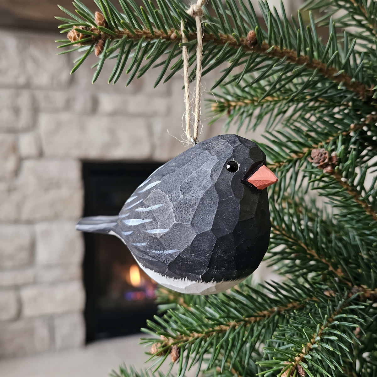 A carved wooden Dark-eyed Junco ornament, shaped like a dark bird with a bright orange beak, hanging from a natural fiber string on a pine tree branch in front of a blurred fireplace background.