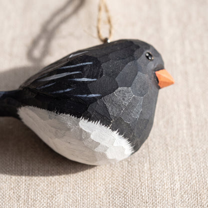 A hand-carved wooden Dark-eyed Junco ornament with a black and white painted finish and a small orange beak, hanging against a beige linen background.