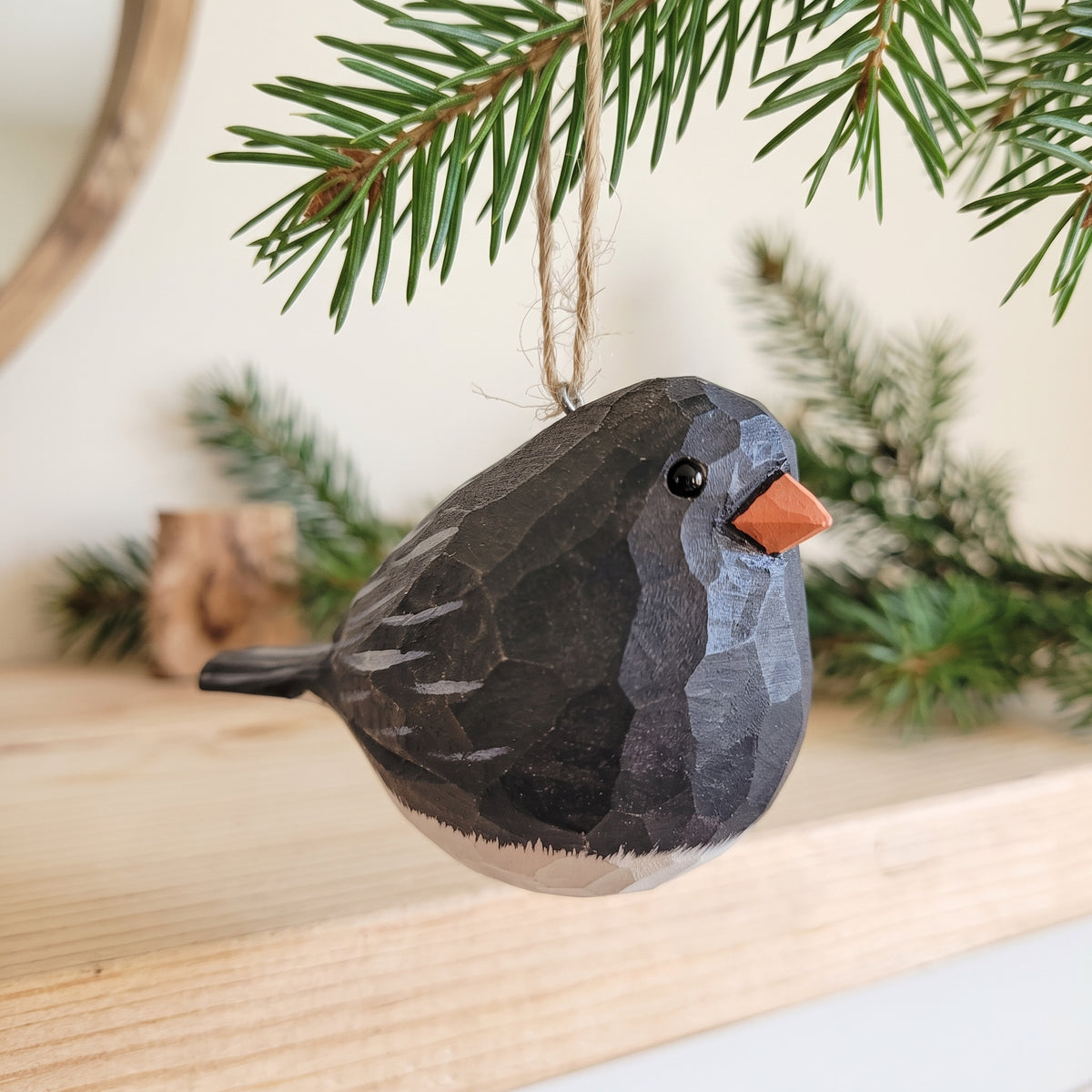 A carved wooden Dark-eyed Junco ornament with a dark gray and black body, white underbelly, and orange beak, hanging from a twine string under evergreen branches.