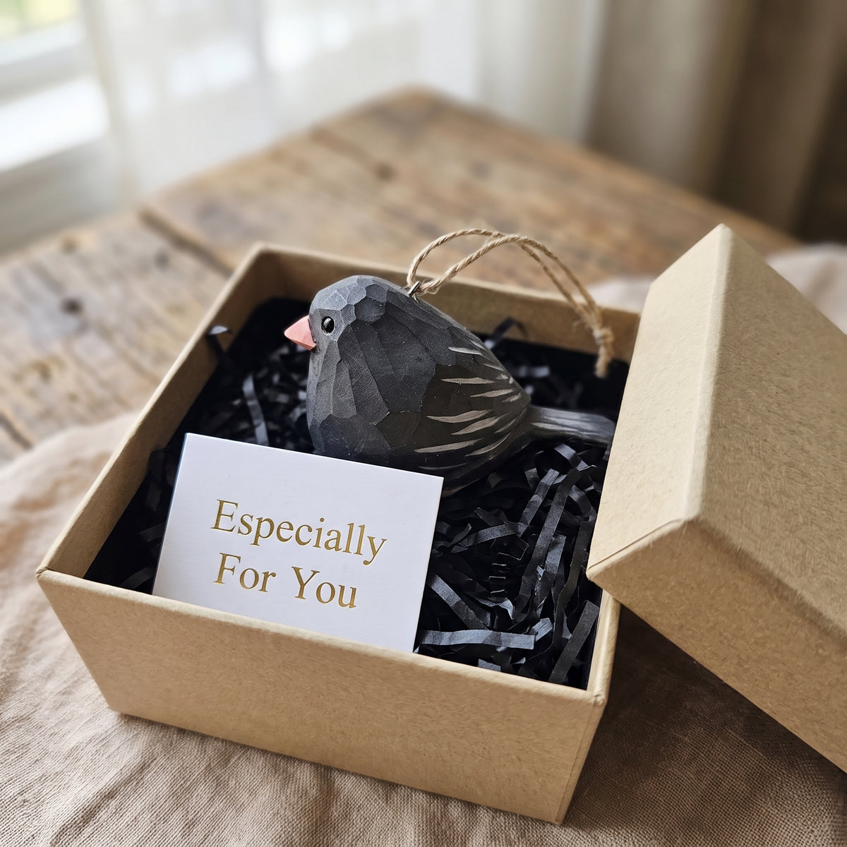 A small Dark-eyed Junco-shaped ornament in dark gray with a pink beak and white tail stripes, presented in a kraft gift box with black shredded paper and a card that reads 'Especially For You'.