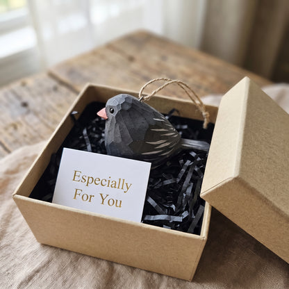A small Dark-eyed Junco-shaped ornament in dark gray with a pink beak and white tail stripes, presented in a kraft gift box with black shredded paper and a card that reads 'Especially For You'.