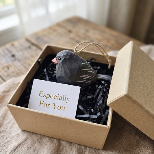 A small Dark-eyed Junco-shaped ornament in dark gray with a pink beak and white tail stripes, presented in a kraft gift box with black shredded paper and a card that reads 'Especially For You'.