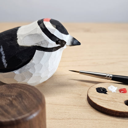 A carved wooden Downy Woodpecker figurine with a black, white, and gray geometric pattern and a small red accent on its head, displayed on a light wood surface next to a paintbrush and a small wooden paint palette.