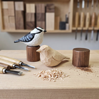Two geometric wooden Downy Woodpecker carvings displayed on a workbench with carving tools and wood shavings. One bird is carved from light natural wood and sits on a cylindrical stand, while the other is carved from darker wood and stands on a larger dark wooden base.