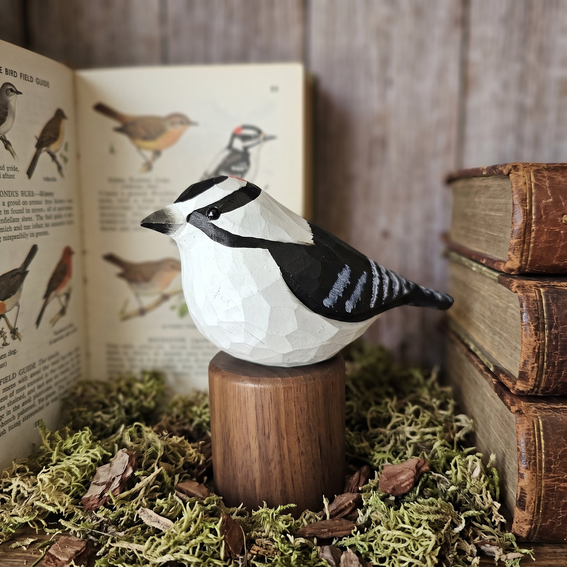 A carved Downy Woodpecker figurine with a geometric, faceted surface design, perched on a cylindrical wooden pedestal. The figurine is styled in a rustic setting with moss, old books, and an open vintage bird field guide against a wooden background.