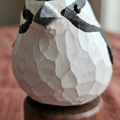 A close-up of a carved wooden Downy Woodpecker figurine with a textured, faceted surface painted in white and black, perched on a round wooden base.