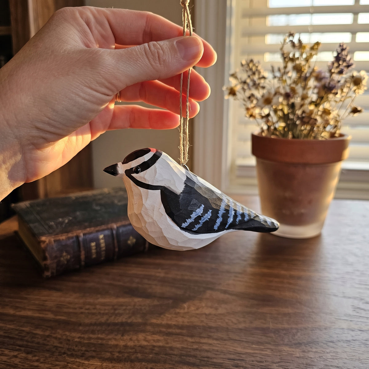 A hand holding up a hand-carved Downy Woodpecker wooden ornament by its jute string against a sunlit window and rustic desk.