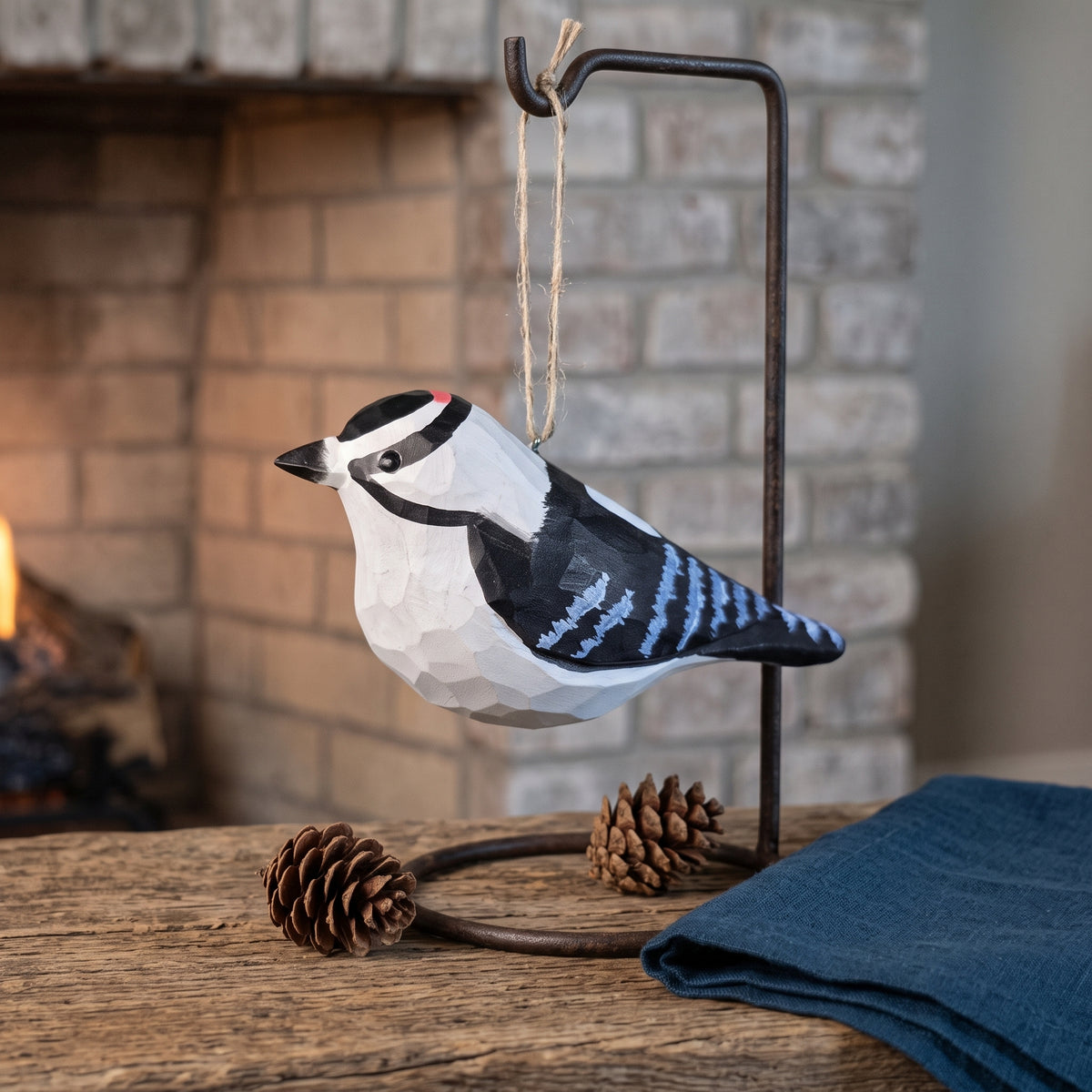 Hand-carved Downy Woodpecker wooden ornament hanging on a black iron display stand against a rustic brick fireplace background.