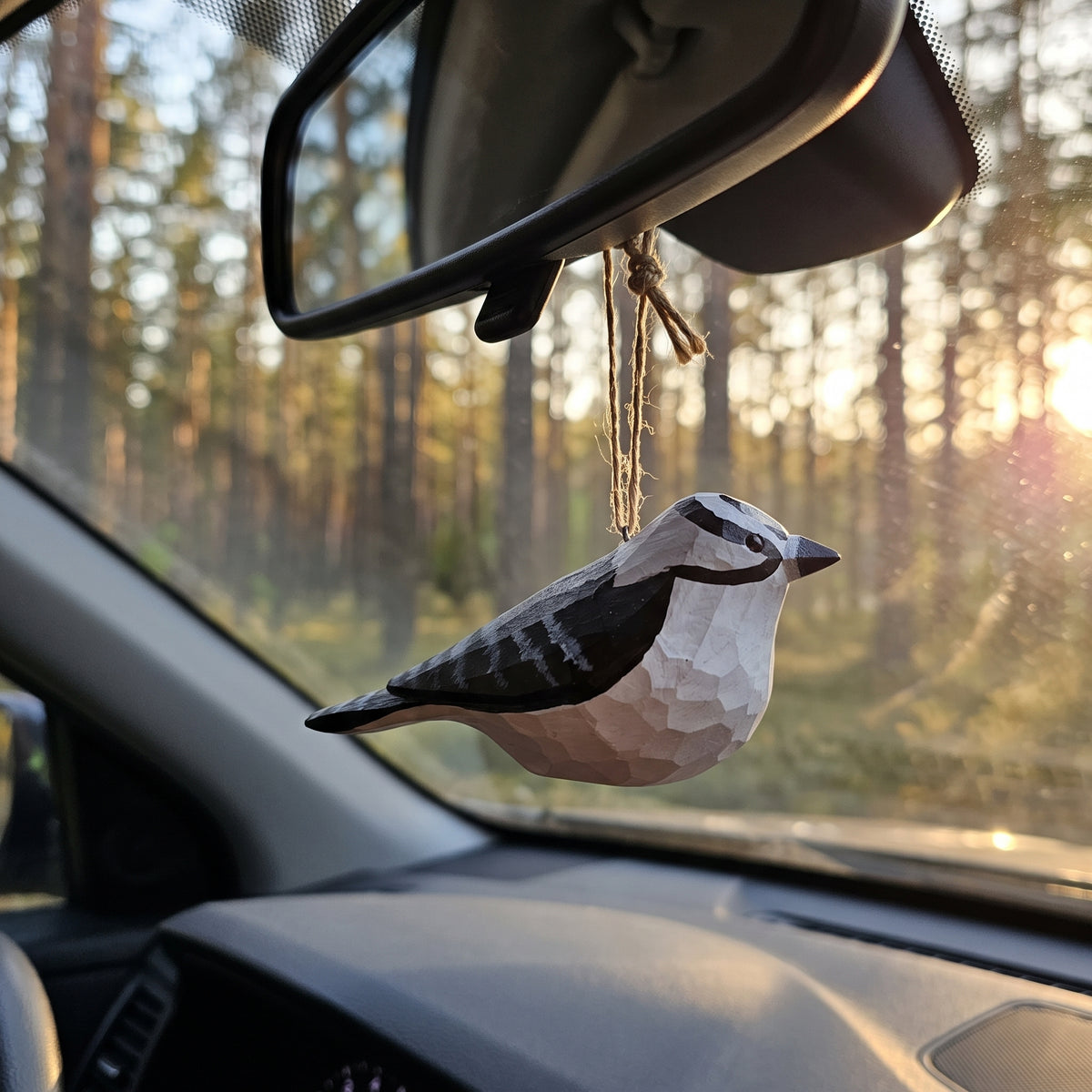 Hand-carved Downy Woodpecker wooden ornament hanging from a car rearview mirror against a sunlit pine forest road.
