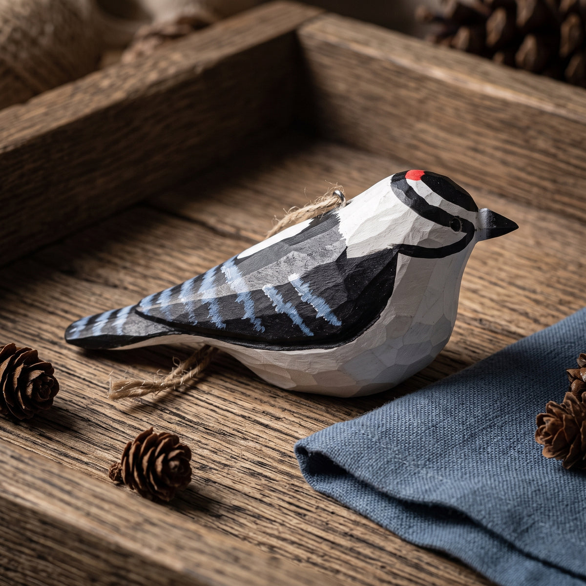 Hand-carved Downy Woodpecker wooden ornament resting on a rustic wooden table next to a blue linen napkin and pinecones.