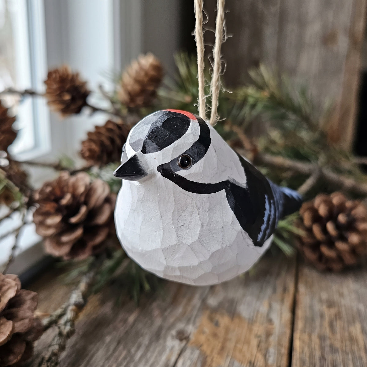 Hand-carved Downy Woodpecker wooden ornament hanging by jute twine on a rustic wooden table with pinecones near a window.