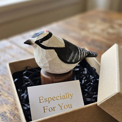 A carved Downy Woodpecker figurine displayed in a kraft gift box with black paper shred and a card that reads 'Especially For You,' set on a rustic wooden table.