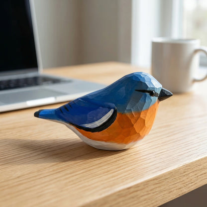 A geometric, low-poly Eastern Bluebird figurine with a blue and orange color scheme sits on a light wood desk in a home office setting, with a laptop and mug blurred in the background.