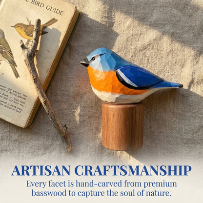 Hand-carved wooden Eastern Bluebird sculpture on a cylindrical wooden base, styled on a linen surface beside an old bird guide book and a small branch. Text at the bottom reads: 'ARTISAN CRAFTSMANSHIP Every facet is hand-carved from premium basswood to capture the soul of nature.