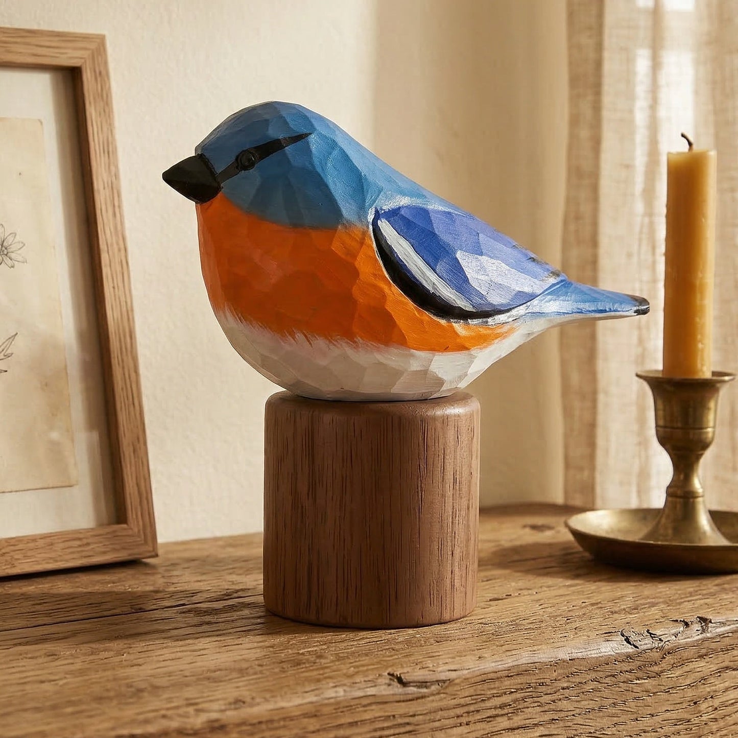 A colorful Eastern Bluebird figurine painted in blue, orange, and white, mounted on a round wooden pedestal on a rustic wooden table. The scene is styled with a framed botanical print and a brass candlestick holder with a lit candle against a warm beige wall and curtain background.