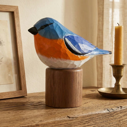 A colorful Eastern Bluebird figurine painted in blue, orange, and white, mounted on a round wooden pedestal on a rustic wooden table. The scene is styled with a framed botanical print and a brass candlestick holder with a lit candle against a warm beige wall and curtain background.